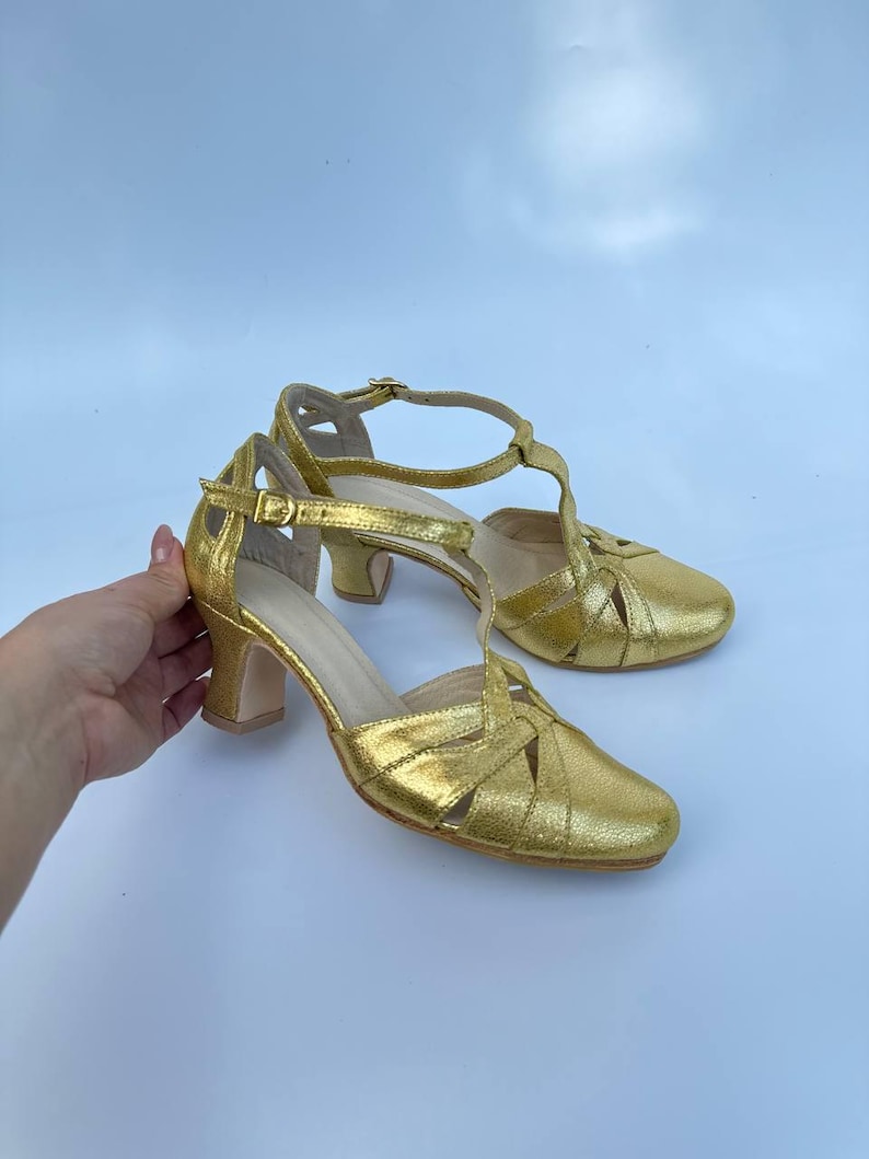 May include: A pair of gold-colored, heeled sandals. The sandals have a woven design across the toes and a T-strap that fastens around the ankle. The heels are approximately 2 inches (5 cm) high.