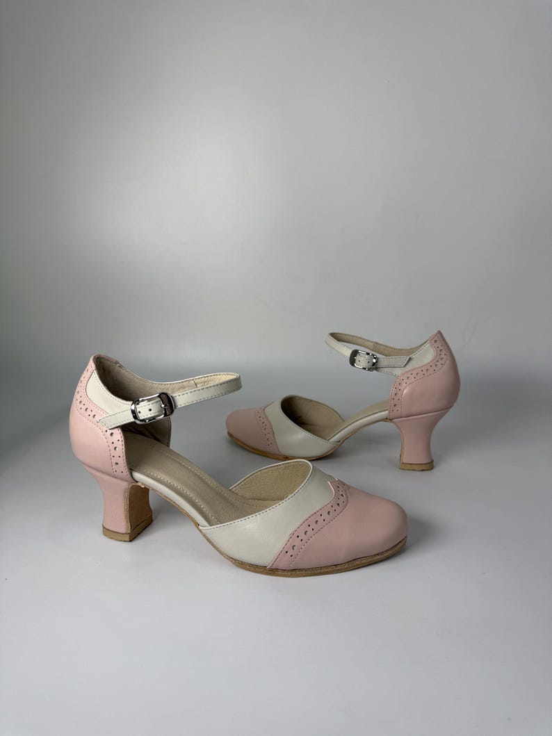 Op de afbeelding: Een paar tweekleurige schoenen met een lichtroze en cr&egrave;mekleurig palet. Deze schoenen hebben een ronde neus, een blokhak van gemiddelde hoogte en een verstelbare enkelband met een zilverkleurige metalen gesp. Het ontwerp bevat decoratieve perforaties in brogue-stijl langs de naden van de neus en de hiel.