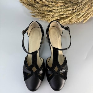 Puede incluir: Un par de zapatos negros con correa en T que presentan una combinaci&oacute;n de texturas lisas y de ante. Los zapatos tienen una puntera redondeada, un dise&ntilde;o recortado en el empeine y una fina correa en el tobillo con una peque&ntilde;a hebilla met&aacute;lica.