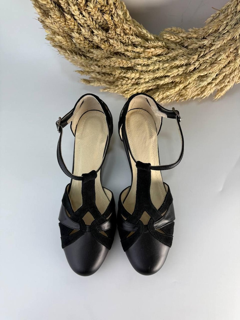 Puede incluir: Un par de zapatos negros con correa en T que presentan una combinaci&oacute;n de texturas lisas y de ante. Los zapatos tienen una puntera redondeada, un dise&ntilde;o recortado en el empeine y una fina correa en el tobillo con una peque&ntilde;a hebilla met&aacute;lica.