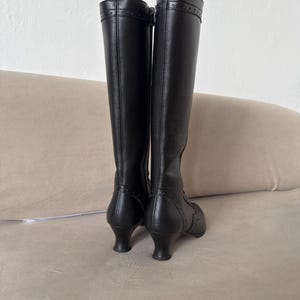 May include: A pair of black knee-high boots featuring a smooth finish, a side zipper closure, and a curved kitten heel. The boots include decorative stitching and small perforated details along the heel counter and the top collar of the shaft.