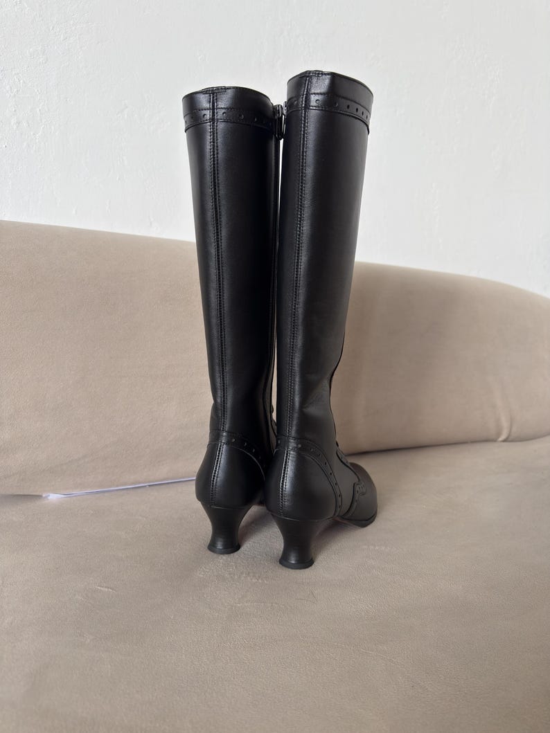 May include: A pair of black knee-high boots featuring a smooth finish, a side zipper closure, and a curved kitten heel. The boots include decorative stitching and small perforated details along the heel counter and the top collar of the shaft.