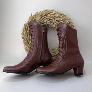 May include: A pair of mid-calf brown boots featuring a lace-up front with dark brown laces, decorative perforations along the seams, a side zipper closure, and a block heel with a black textured sole. The boots have a smooth finish.