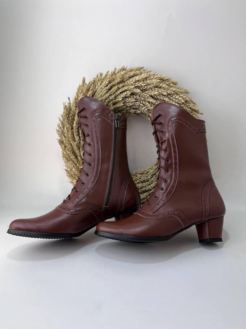 May include: A pair of mid-calf brown boots featuring a lace-up front with dark brown laces, decorative perforations along the seams, a side zipper closure, and a block heel with a black textured sole. The boots have a smooth finish.