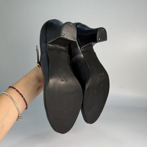 May include: Black leather heeled shoes with a strap and buckle closure. The shoes have a rounded toe and a block heel. The soles are dark and appear to be made of a durable material. The shoes are shown from the bottom up.