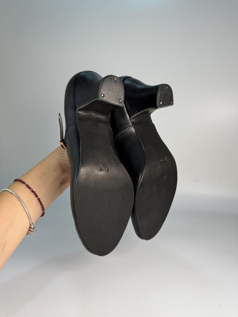 May include: Black leather heeled shoes with a strap and buckle closure. The shoes have a rounded toe and a block heel. The soles are dark and appear to be made of a durable material. The shoes are shown from the bottom up.