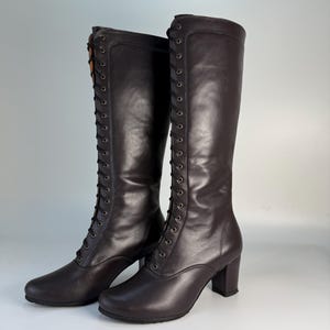 May include: A pair of dark brown knee-high boots with a lace-up design. The boots have a rounded toe, a block heel, and a smooth leather texture. The laces are black, and the boots are set against a plain background.