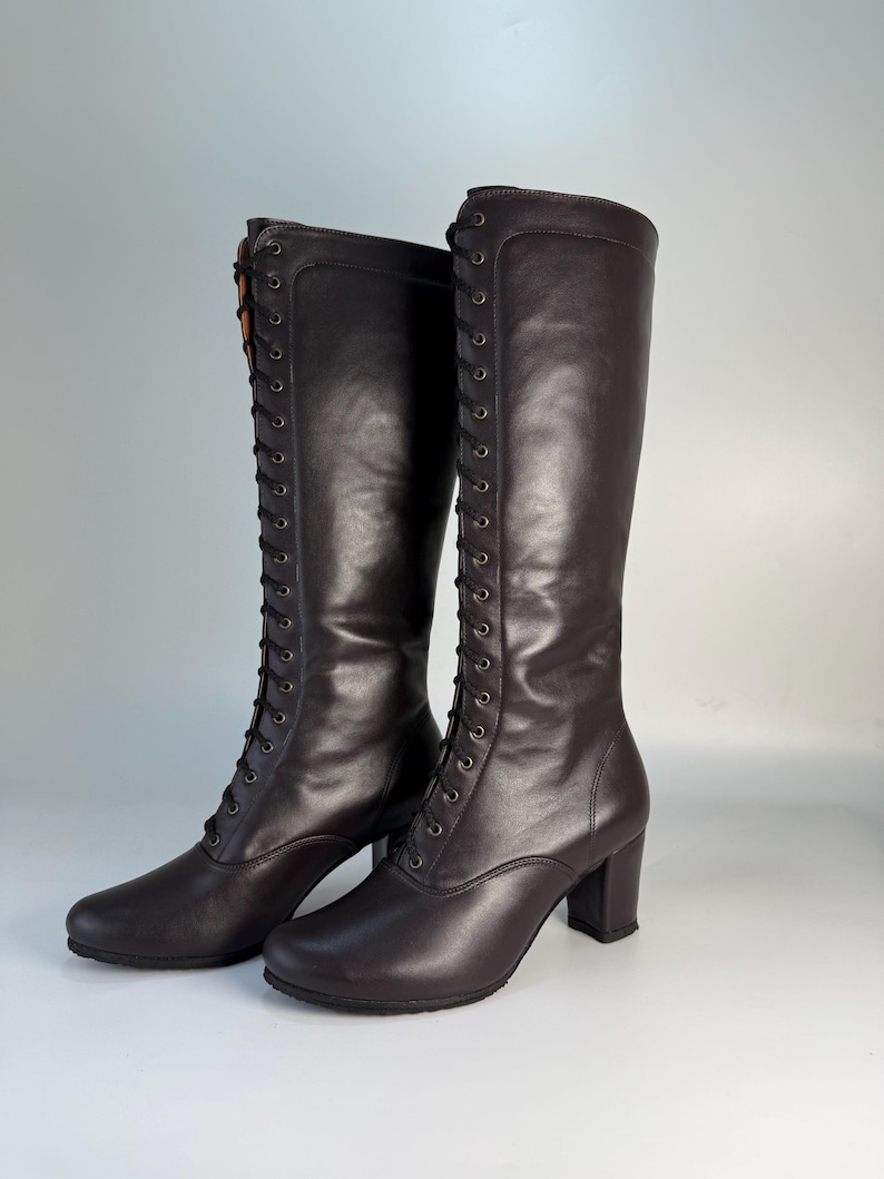 May include: A pair of dark brown knee-high boots with a lace-up design. The boots have a rounded toe, a block heel, and a smooth leather texture. The laces are black, and the boots are set against a plain background.