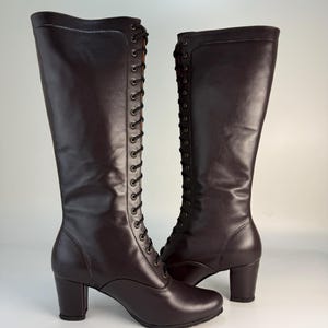 May include: A pair of knee-high, dark brown leather boots with a lace-up design. The boots have a rounded toe and a block heel. The laces are black, and the boots have a smooth, polished finish.