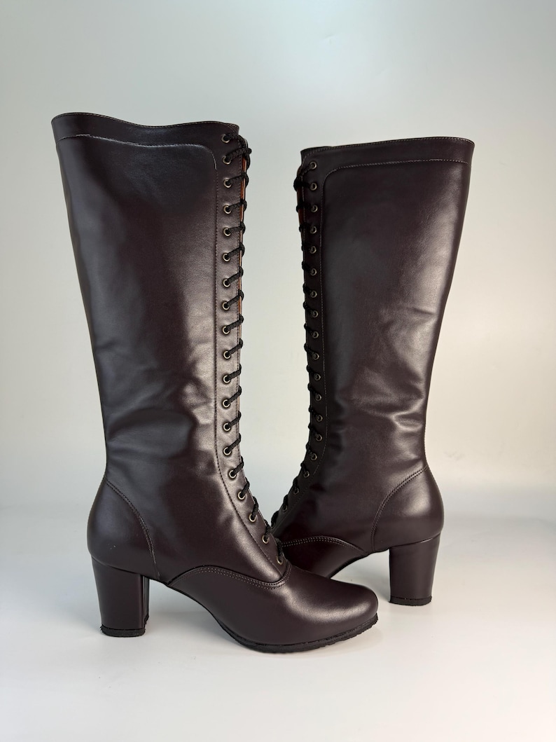 May include: A pair of knee-high, dark brown leather boots with a lace-up design. The boots have a rounded toe and a block heel. The laces are black, and the boots have a smooth, polished finish.