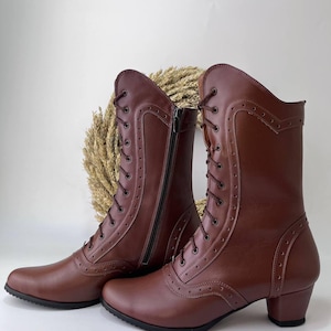 May include: A pair of mid-calf brown boots featuring a lace-up front with metal eyelets, decorative brogue-style perforated detailing along the seams, a side zipper closure, and a low block heel with a black rubber sole.