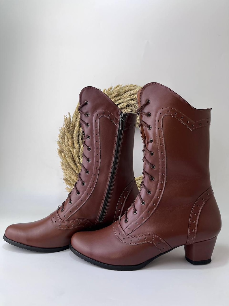 May include: A pair of mid-calf brown boots featuring a lace-up front with metal eyelets, decorative brogue-style perforated detailing along the seams, a side zipper closure, and a low block heel with a black rubber sole.