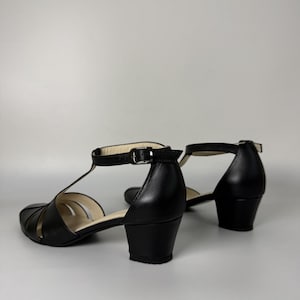 May include: A pair of black leather T-strap sandals featuring a closed toe, a low block heel, and an adjustable ankle strap with a silver-toned metal buckle. The interior lining is a light beige colour, and the soles are black.