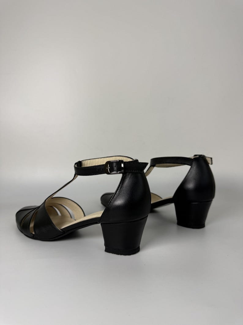 May include: A pair of black leather T-strap sandals featuring a closed toe, a low block heel, and an adjustable ankle strap with a silver-toned metal buckle. The interior lining is a light beige colour, and the soles are black.