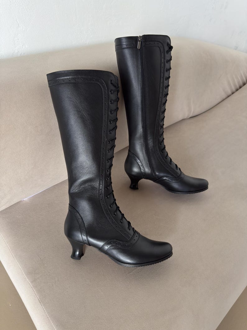 May include: A pair of black knee-high boots featuring a front lace-up design with thin black laces, decorative brogue-style perforations along the seams, a rounded toe, and a low, flared kitten heel. Each boot includes a side zipper closure.