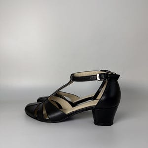 May include: A pair of black T-strap shoes with a rounded toe, cut-out side panels, and a low block heel. The shoes feature a thin ankle strap with a silver-toned metal buckle closure and a light-coloured interior lining visible at the heel and footbed.