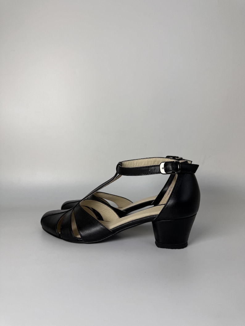 May include: A pair of black T-strap shoes with a rounded toe, cut-out side panels, and a low block heel. The shoes feature a thin ankle strap with a silver-toned metal buckle closure and a light-coloured interior lining visible at the heel and footbed.
