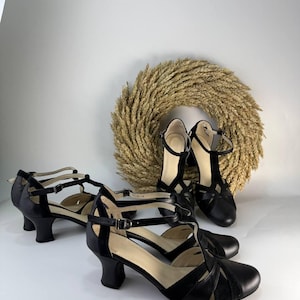Puede incluir: Un par de zapatos de tac&oacute;n negros con tira en T, punta redondeada, tac&oacute;n de bloque de altura media y correa ajustable al tobillo con hebilla met&aacute;lica. Los zapatos est&aacute;n confeccionados con paneles de cuero negro liso y texturizado.