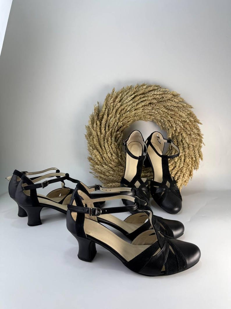 Puede incluir: Un par de zapatos de tac&oacute;n negros con tira en T, punta redondeada, tac&oacute;n de bloque de altura media y correa ajustable al tobillo con hebilla met&aacute;lica. Los zapatos est&aacute;n confeccionados con paneles de cuero negro liso y texturizado.
