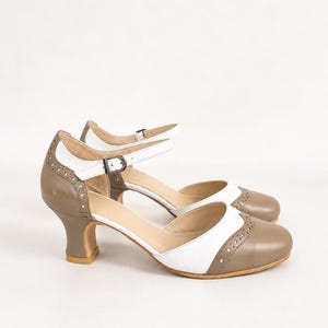 May include: A pair of two-tone Mary Jane heels featuring a taupe and white color scheme. The shoes have a rounded toe, a cutout side design, and a thin ankle strap with a metal buckle. Decorative brogue-style perforations line the edges.