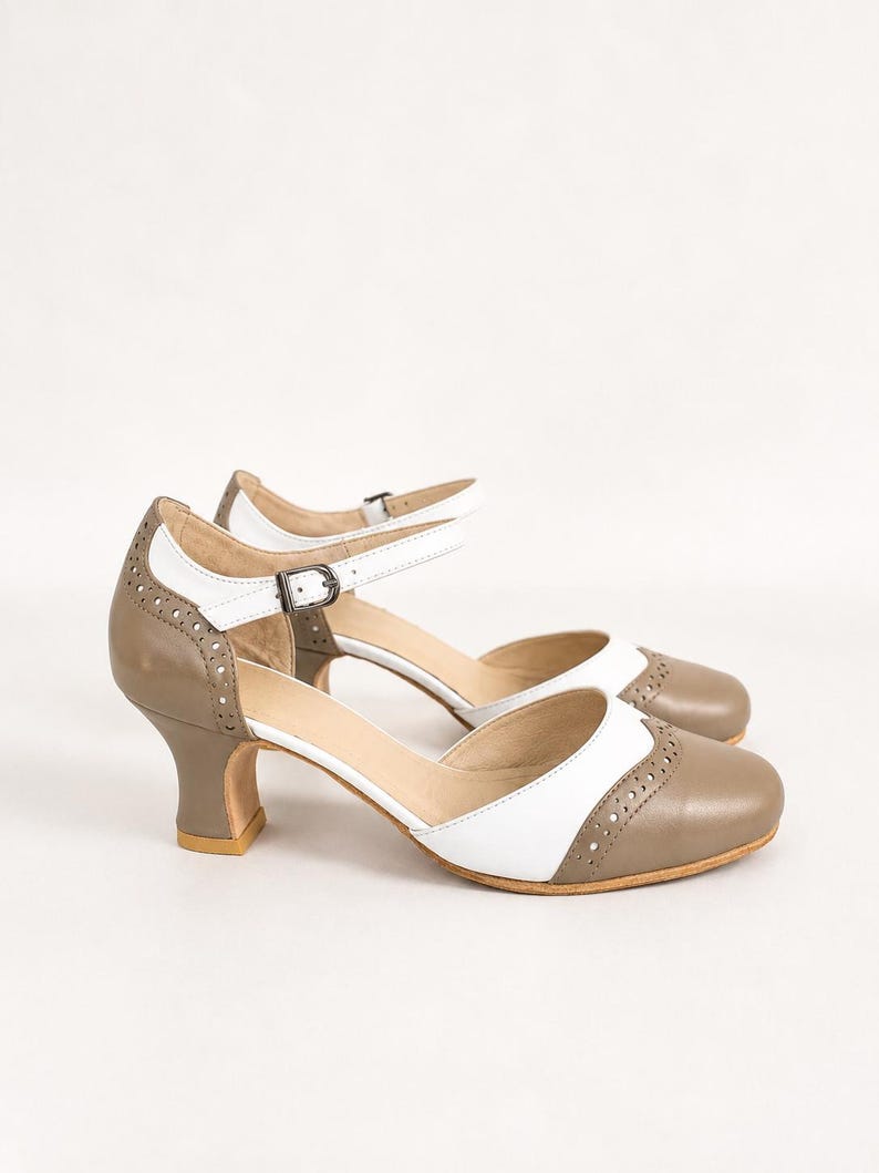 May include: A pair of two-tone Mary Jane heels featuring a taupe and white color scheme. The shoes have a rounded toe, a cutout side design, and a thin ankle strap with a metal buckle. Decorative brogue-style perforations line the edges.