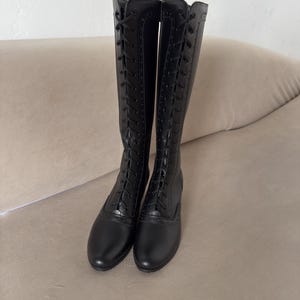 May include: A pair of knee-high black boots featuring a full-length lace-up front closure with black laces and eyelets. The boots have a rounded toe, a low heel, and decorative brogue-style perforated detailing along the lace panels.