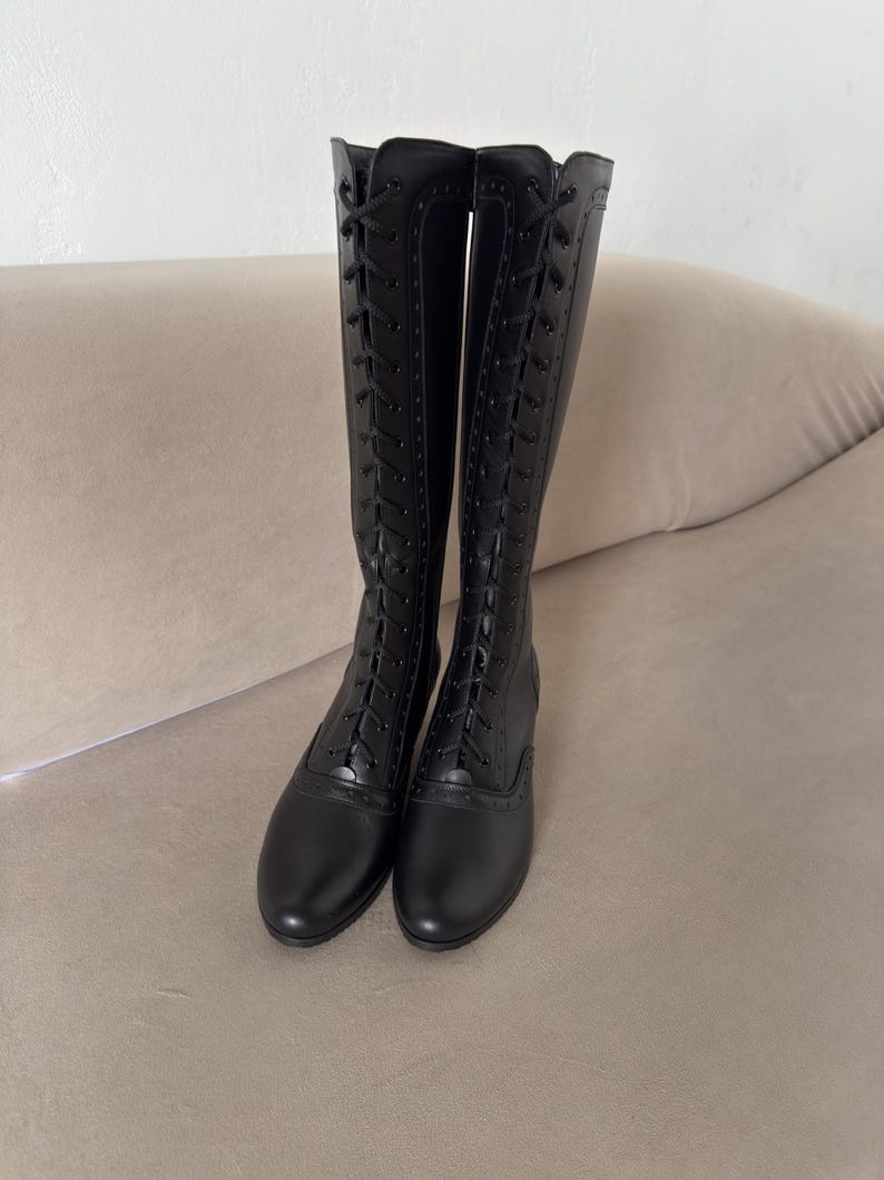 May include: A pair of knee-high black boots featuring a full-length lace-up front closure with black laces and eyelets. The boots have a rounded toe, a low heel, and decorative brogue-style perforated detailing along the lace panels.