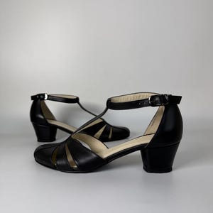 May include: A pair of black leather T-strap shoes featuring a rounded toe, a low block heel, and an adjustable ankle strap with a silver-toned metal buckle. The upper part of the shoe has a series of cutouts creating a fan-like pattern.