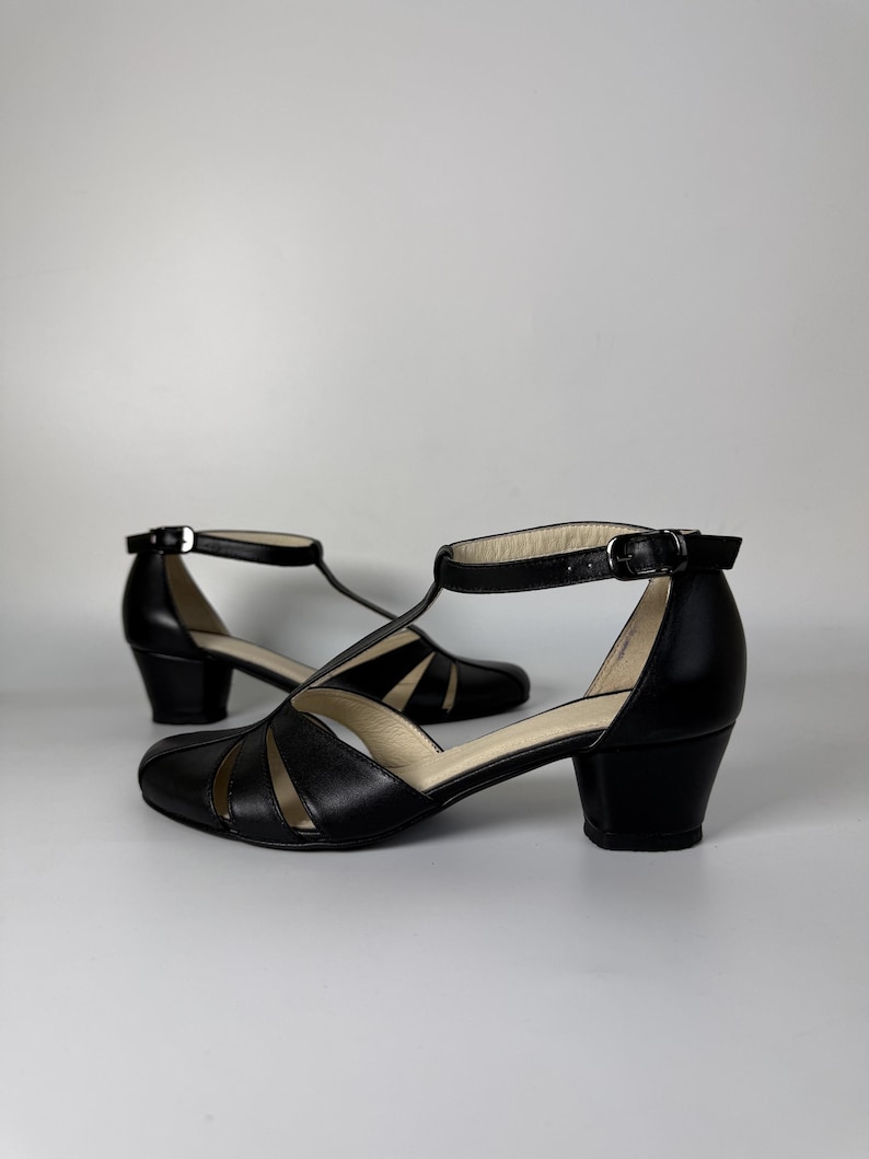 May include: A pair of black leather T-strap shoes featuring a rounded toe, a low block heel, and an adjustable ankle strap with a silver-toned metal buckle. The upper part of the shoe has a series of cutouts creating a fan-like pattern.