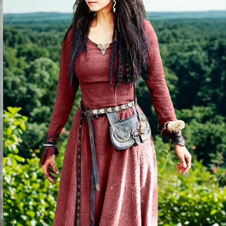 May include: A long-sleeved, burgundy-colored dress with a scoop neckline and a decorative pendant. The dress is cinched at the waist with a leather belt and a small pouch. The sleeves have decorative cuffs. The background is a blurred green forest.