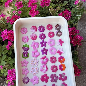 Peut inclure: Un plateau blanc rempli d'une variété d'ornements en forme de fleurs aux couleurs vives. Les fleurs sont roses, violettes et oranges, avec des paillettes et des motifs marbrés. Le plateau est posé devant des fleurs roses.