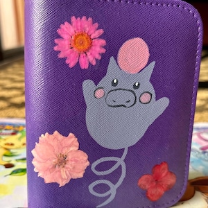 May include: A purple cardholder decorated with a gray cartoon character and pressed flowers. The cardholder has a stitched border and features a pink and orange flower, a pink flower, and a red flower.