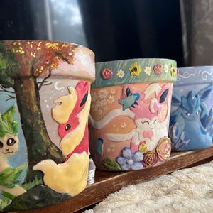 May include: A set of four hand-painted ceramic flower pots. Each pot features a different colorful design of cartoon characters and nature scenes. The pots are displayed on a wooden shelf.