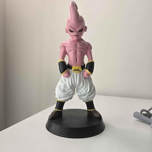 May include: A pink figurine of a character with a distinctive antenna-like head, stern expression, and muscular physique. The figure wears white pants, black armbands, and boots, with a gold belt buckle. It stands on a black circular base.