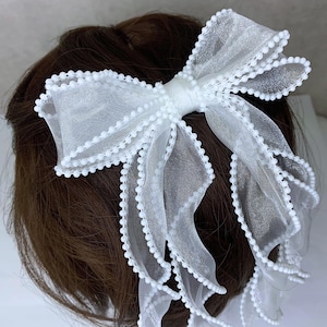 Op de afbeelding: Een witte haarstrik van doorschijnende stof, met een gelaagd ontwerp. De randen zijn afgewerkt met kleine, witte kralen. De strik is vastgemaakt met een bijpassend wit lint. Een stijlvol haaraccessoire voor speciale gelegenheden.