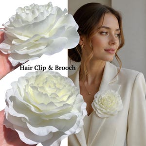 May include: A white flower hair clip and brooch featuring layered, ruffled petals. The accessory is shown held in a hand and pinned to a cream-colored blazer. The text "Hair Clip & Brooch" is printed in black in the center of the image.