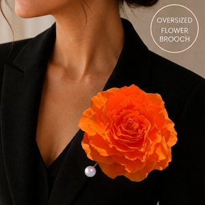 May include: An oversized orange flower brooch with a pearl pin is attached to a black blazer. The brooch is a large, vibrant orange, resembling a rose. The text "OVERSIZED FLOWER BROOCH" is in a white circle.