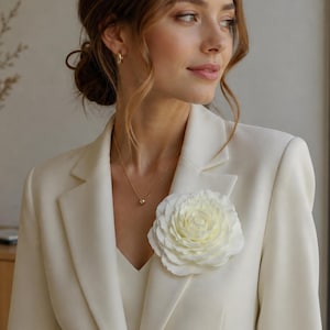 May include: A large, off-white fabric flower brooch pinned to the lapel of a cream-colored blazer. The flower features layered, rounded petals in a soft, matte finish. The blazer has a structured collar and a V-neckline, paired with a gold necklace.