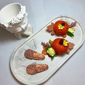 Può includere: Due fermagli per capelli con zucche in feltro arancione, fiori gialli e foglie marroni glitterate, insieme a due fermagli rettangolari ricoperti di paillettes quadrate color rame, su un vassoio ovale bianco con motivi a farfalla.
