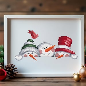 May include: Cross-stitch artwork in a white frame featuring three snowmen with carrot noses and winter hats. A red bird flies above. The snowmen are smiling. The artwork is displayed on a wooden surface with Christmas decorations.