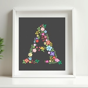 Puede incluir: Una obra de arte enmarcada presenta una "A" floral sobre un fondo gris oscuro. La letra está compuesta por coloridas flores bordadas en tonos de rojo, amarillo, azul y morado. La obra se exhibe en un marco blanco.