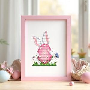 May include: A framed cross-stitch artwork featuring a pink Easter egg with bunny ears and feet, set on green grass. The artwork is in a pink frame, with Easter eggs and decorations in the foreground.