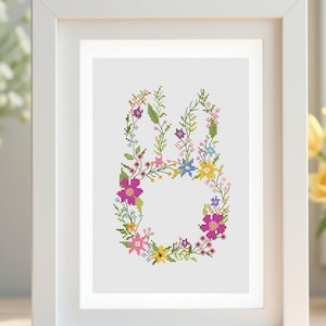 Peut inclure: Broderie au point de croix d'un lapin formé de fleurs et de verdure colorées, encadrée en blanc. Le motif floral comprend des éléments roses, jaunes, bleus et verts, créant un thème printanier ou pascal. L'œuvre est encadrée dans un cadre blanc.