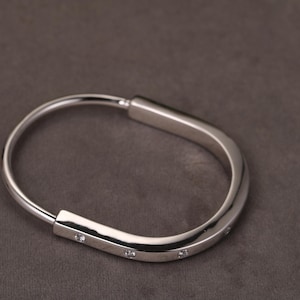 May include: A silver bracelet with a curved, oval shape. The bracelet has small, clear gemstones set along the straight edge. The jewellery is displayed on a dark grey background.