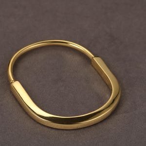 May include: A gold-coloured bracelet with a curved, oval design. The bracelet has a smooth, polished finish and a minimalist aesthetic. The bracelet is approximately 6.35 cm in diameter and is a piece of jewellery.
