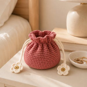 Könnte beinhalten: Ein gehäkelter Beutel mit Kordelzug in Altrosa mit strukturiertem Maschenbild. Die Tasche verfügt über zwei cremefarbene geflochtene Kordeln zum Zuziehen, die jeweils mit einer kleinen, flachen, cremefarbenen Häkelblume mit gelber Mitte enden.