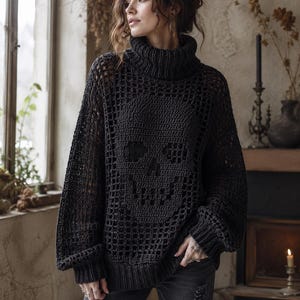 Könnte beinhalten: Ein anthrazitfarbener Langarmpullover mit einem markanten Häkel-Totenkopf-Design auf der Vorderseite. Das Kleidungsstück hat einen hohen Rollkragen, gerippte Bündchen und einen gerippten Saum sowie ein offenes Strickgittermuster.