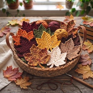 May include: A round woven basket with leather handles containing multiple crocheted maple leaves in various autumn colours including red, orange, yellow, brown, and cream, alongside three balls of yarn in orange, burgundy, and mustard yellow.
