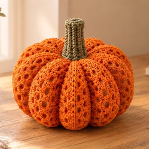 May include: A round, crocheted pumpkin in a vibrant orange hue, featuring a textured, open-work stitch pattern throughout its body. The pumpkin is topped with a short, cylindrical stem crocheted in a muted olive green yarn with a ribbed texture.