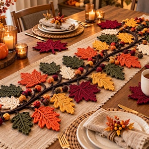 Pode incluir: Um caminho de mesa longo e retangular de tecido bege decorado com uma guirlanda central de crochê tridimensional, apresentando folhas de bordo em tons de vermelho, laranja, amarelo, verde e creme, intercaladas com pequenas bolotas de crochê.